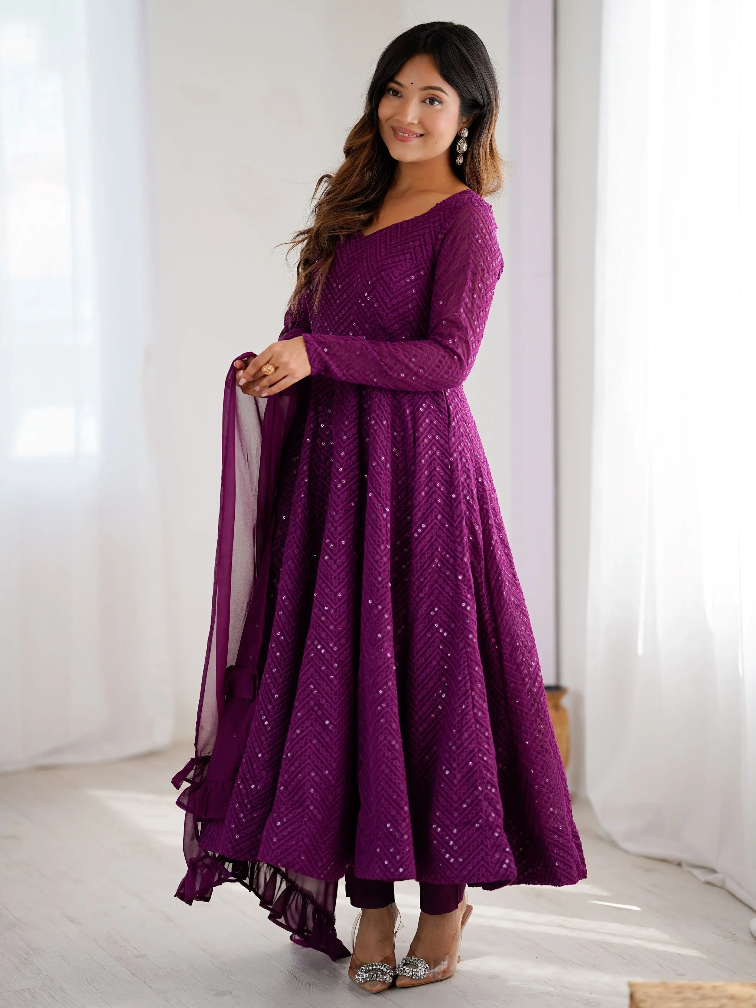 Woman wearing a purple dress with a sheer dupatta against a white curtain background